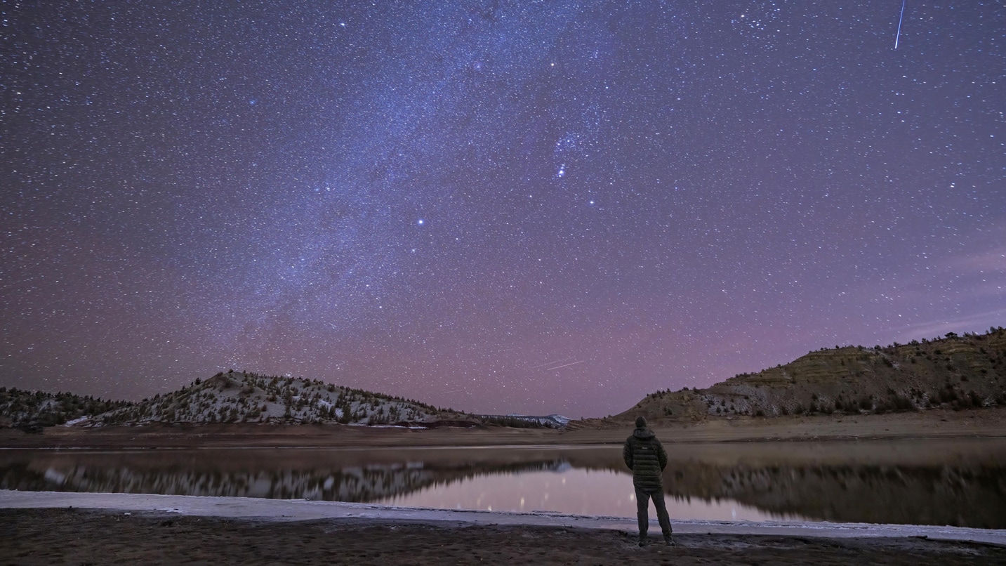Person stands in front of a body of water under a starry night sky.
