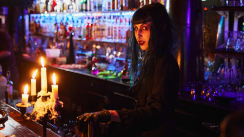 A female bartender in gothic attire and makeup behind a dark, gothic themed bar counter.