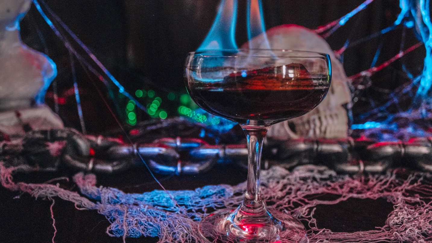 A cocktail is set aflame against a backdrop of gothic and spooky props, including a skull and spiderwebs.
