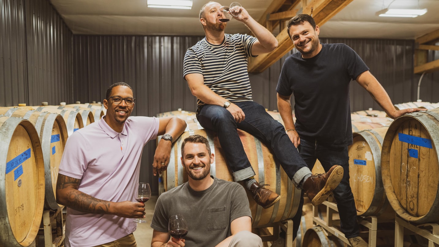 A group of men pose in front of barrels of wine.