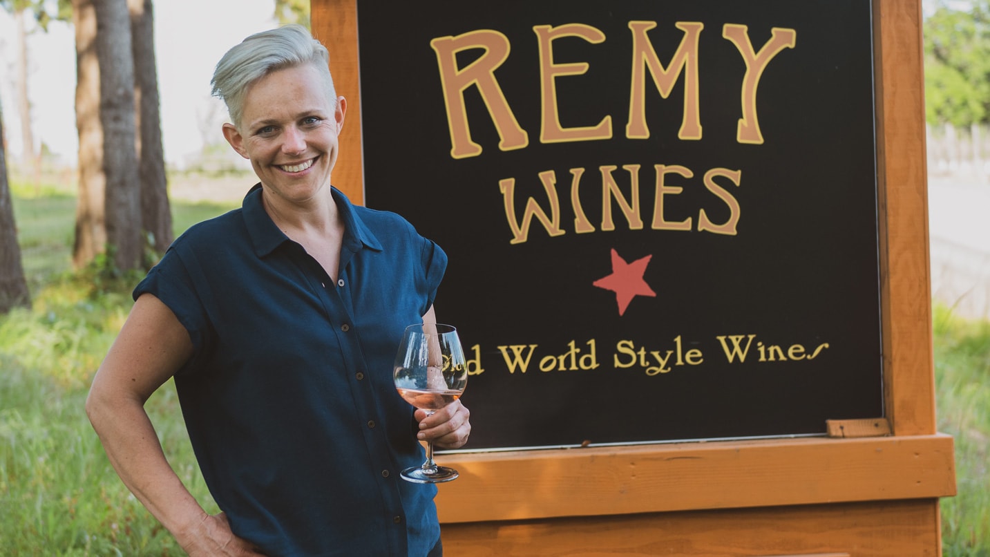 A woman holding a glass of wine in front of a sign for Remy Wines. The sign also reads "Old World Style Wines"