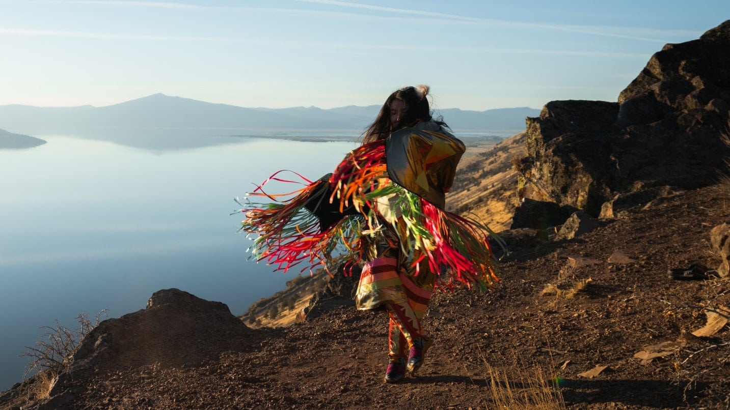 A tribal dancer wears regalia on a dirt trail in front of a lake