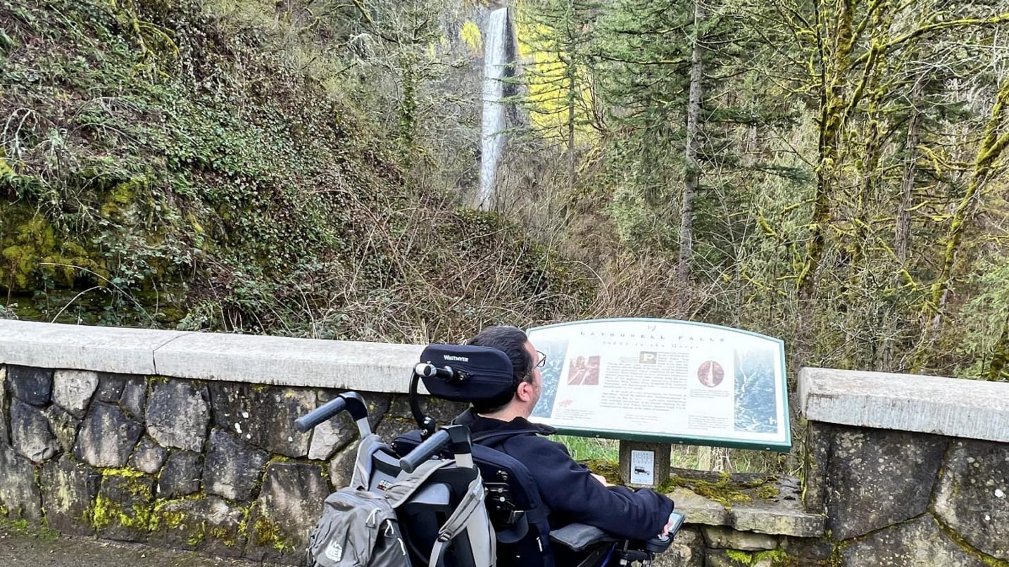 man in wheelchair looks at sign and tall waterfall