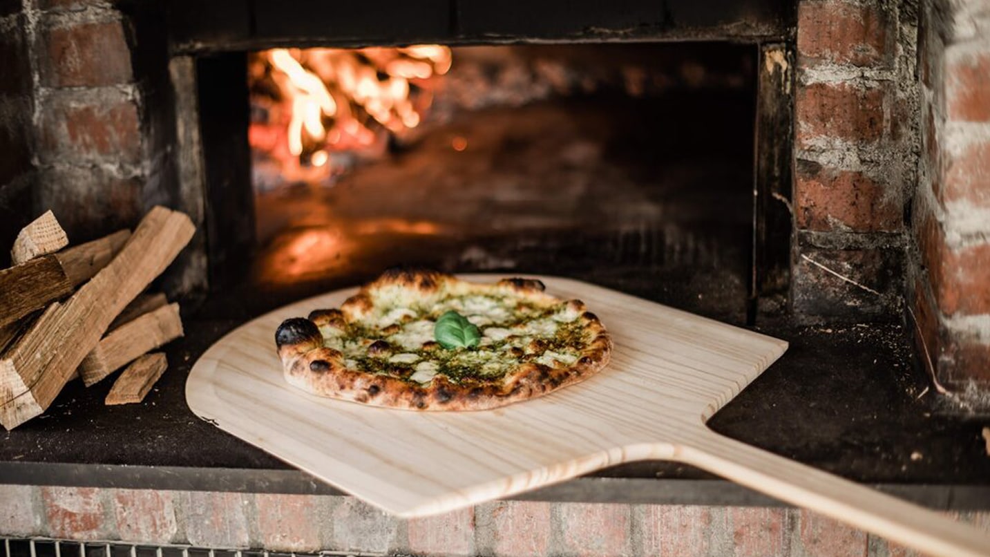 A pesto basil pizza is taken out of a wood-fired oven.