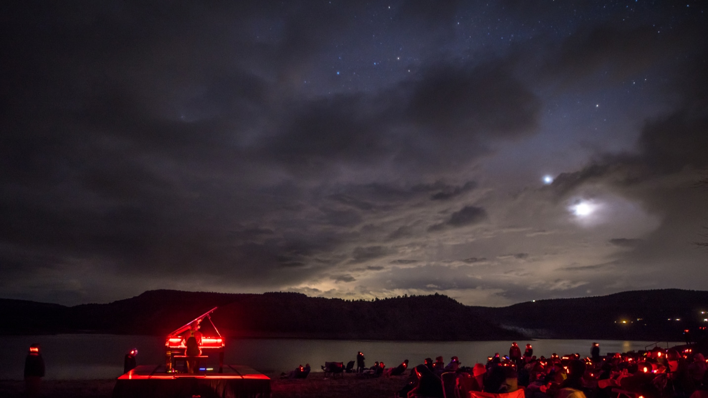 A piano is lit up in red light against a dark starry sky and lake as people sit and watch
