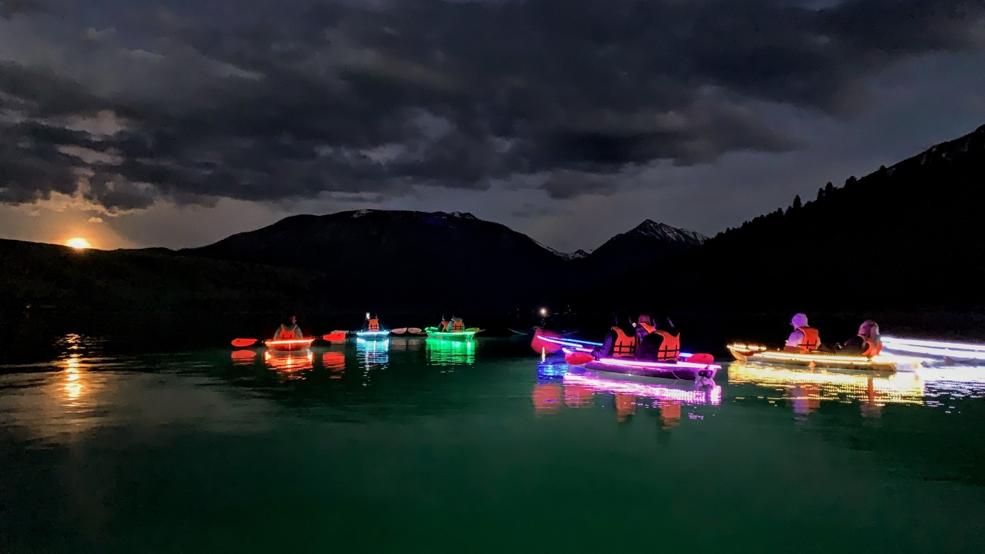 People sit in multi-colored kayaks on a lake with dark sky, moon and mountains in background