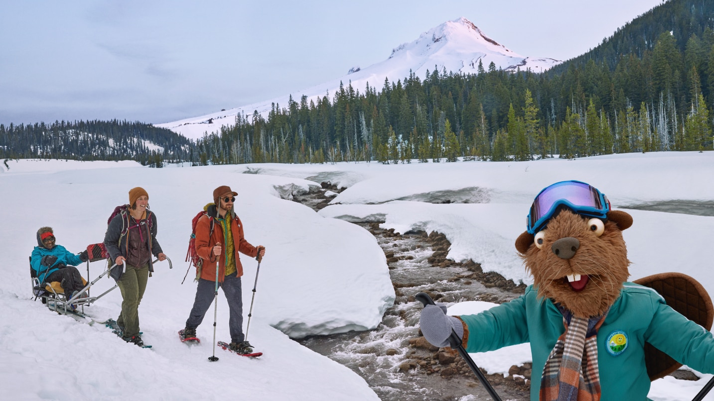 Prominent: a puppet beaver in snow gear. In the background, two trip leaders trek through snow with a person in an adaptive skiing sled.