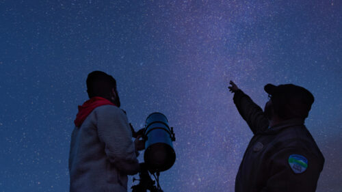 Two people gazing up to a strip of bright stars in the night sky.
