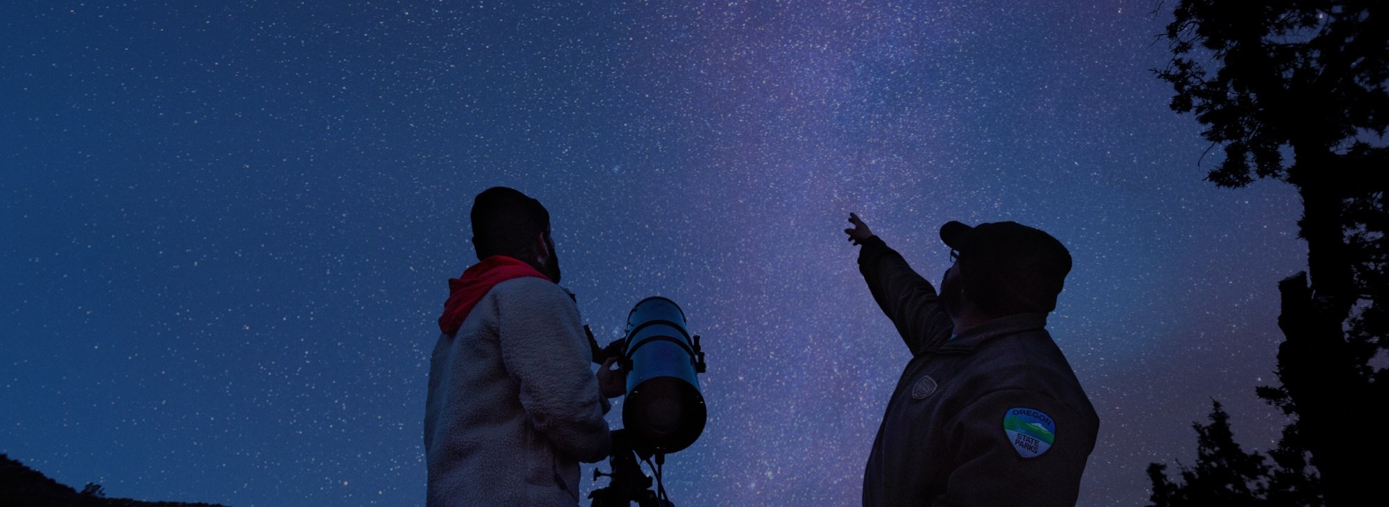 Two people gazing up to a strip of bright stars in the night sky.
