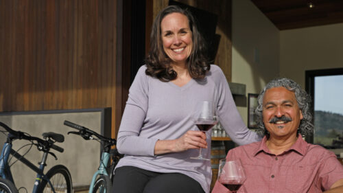 A couple sitting holding wine glasses smile for a portrait. Bicycles can be seen in the background.