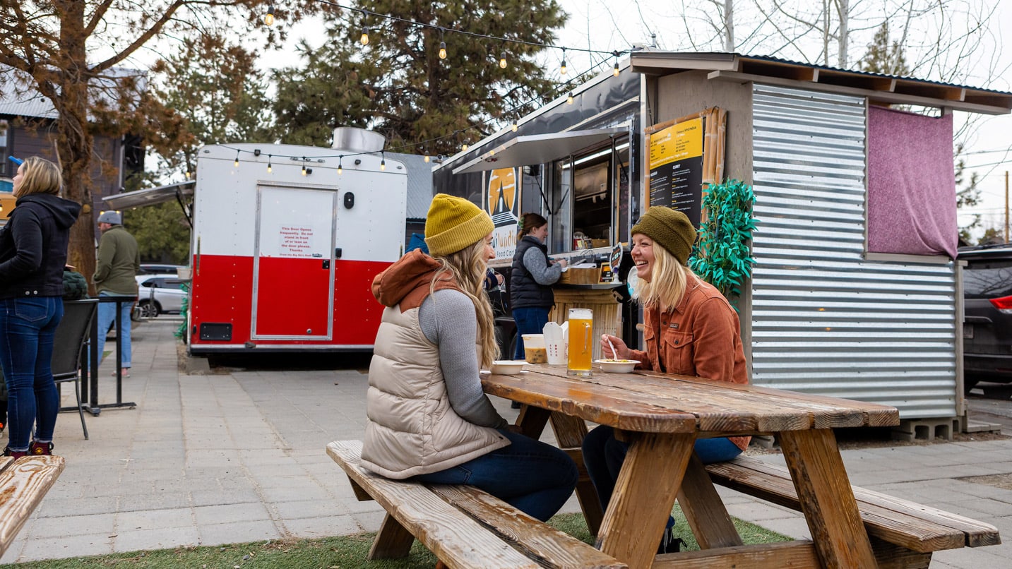 Two women sit at a picnic table in a food cart pod.