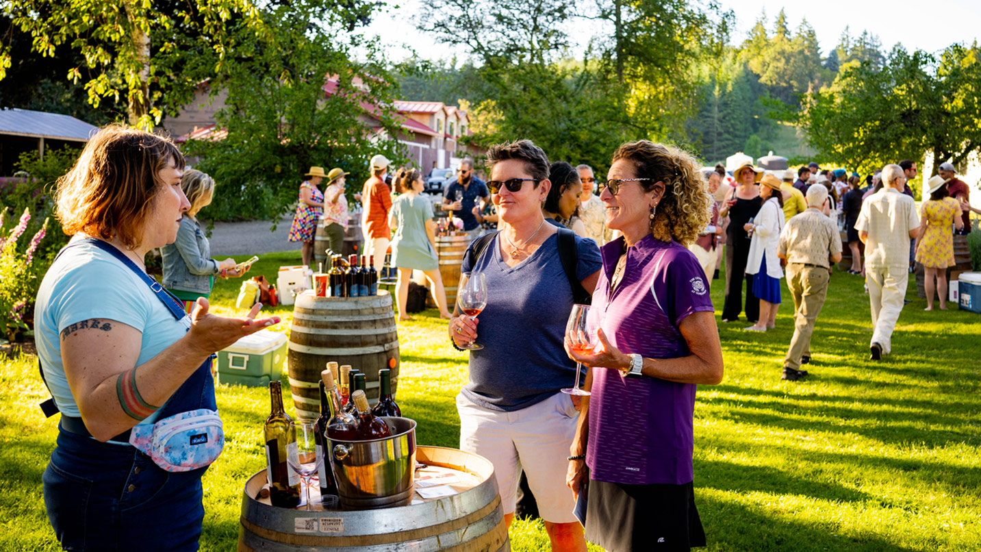 Two women hold wine glasses at lawn gathering with other people in background