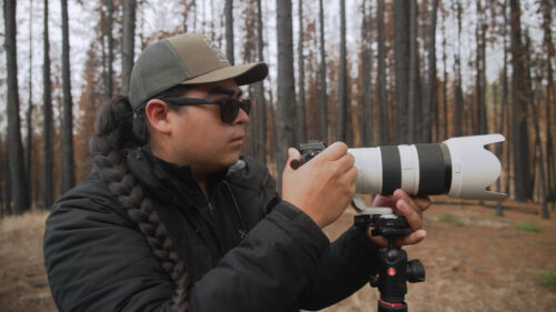man with long braid wears hat and holds a long-lens camera in front of forest