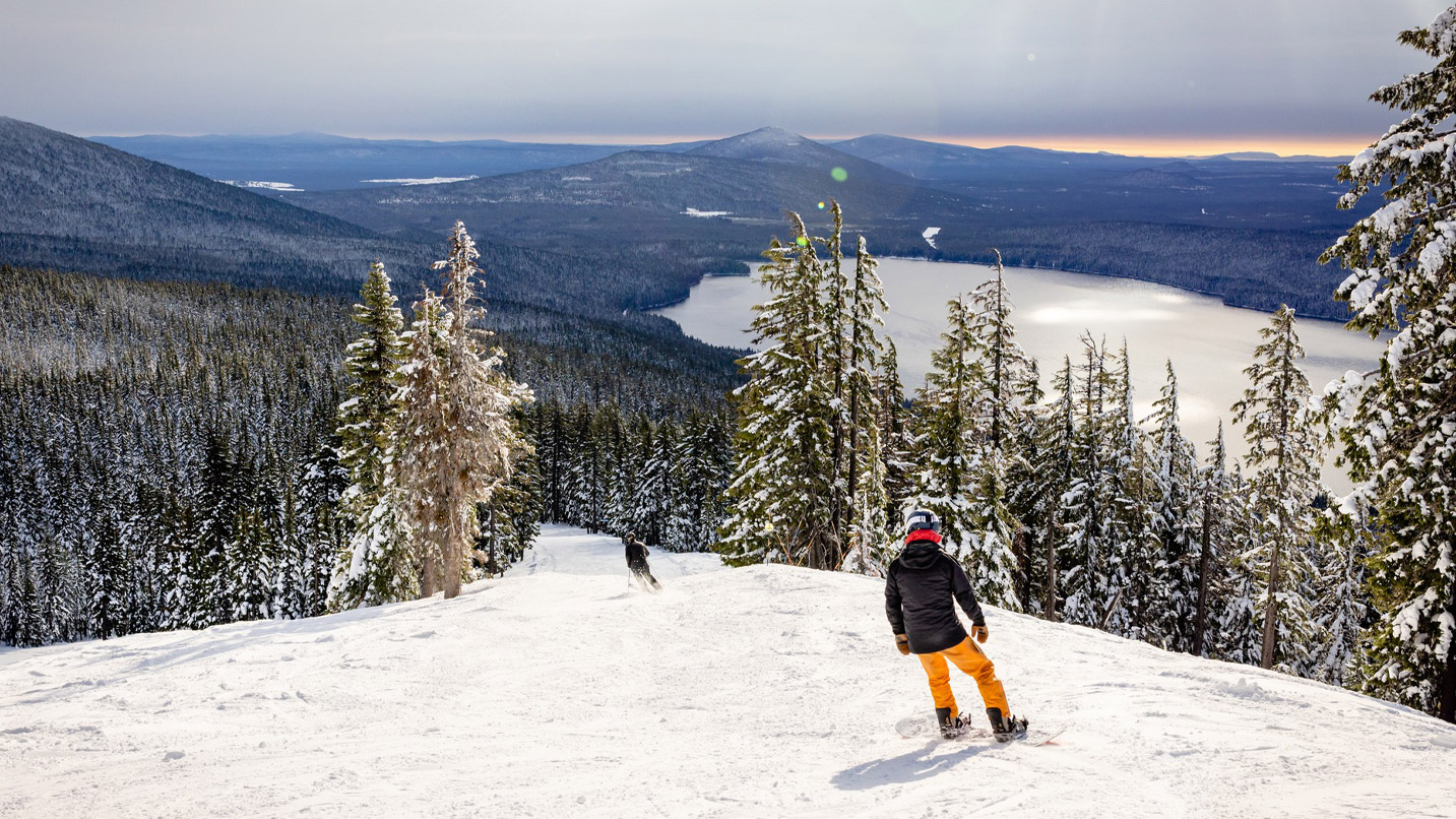 Snowboarder heading down a run with a view of forest and lake below.