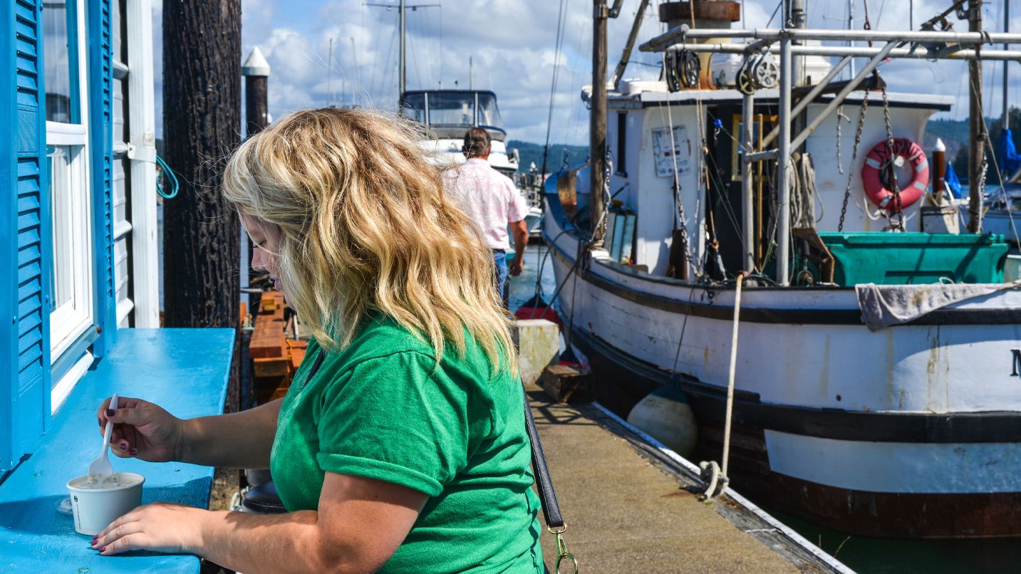A woman enjoys a cup of chowder on an outdoor counter nearby fishing boats.