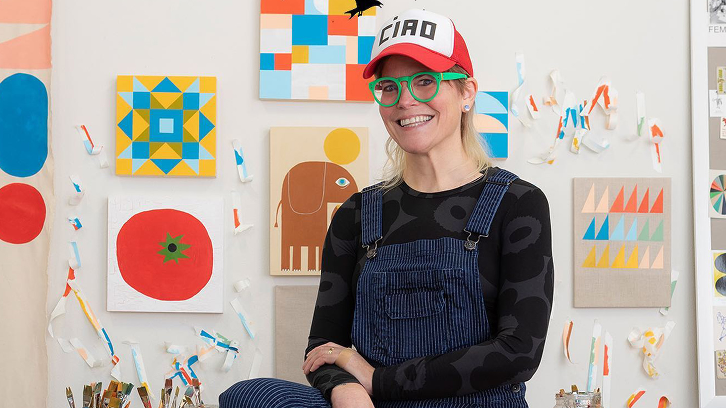 Artist Lisa Cogdon poses in front of a few of her prints in the studio. She is a caucasian woman wearing overalls, bright green glasses and a hat that says "ciao."