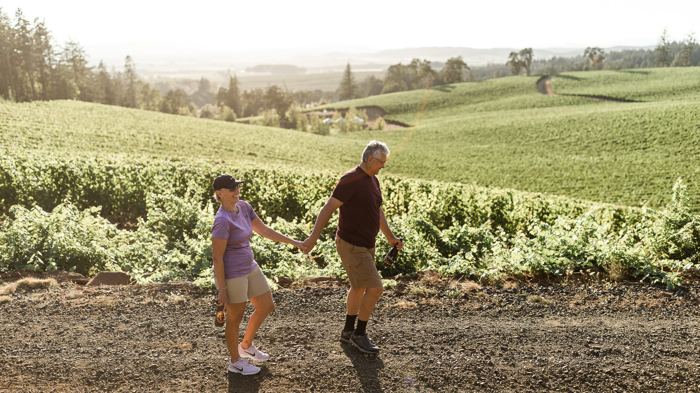 A couple holding hands walking among the hills of a vineyard.