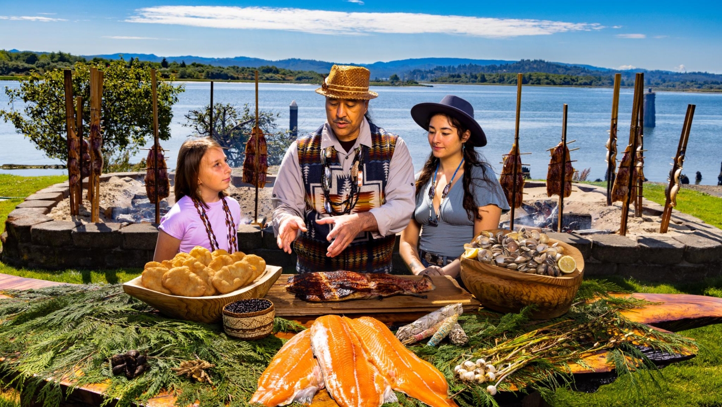 Girl, Man and young woman stand in front of body of water looking at a spread of fresh foods including salmon, berries and clams.