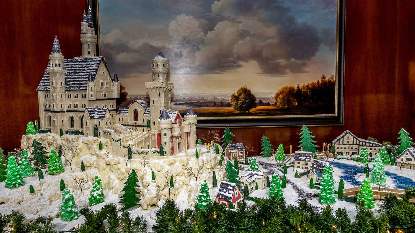 Elaborate gingerbread village, consisting of a large castle structure on a rocky cliff overlooking a small village of homes, a lake, a bridge and trees.