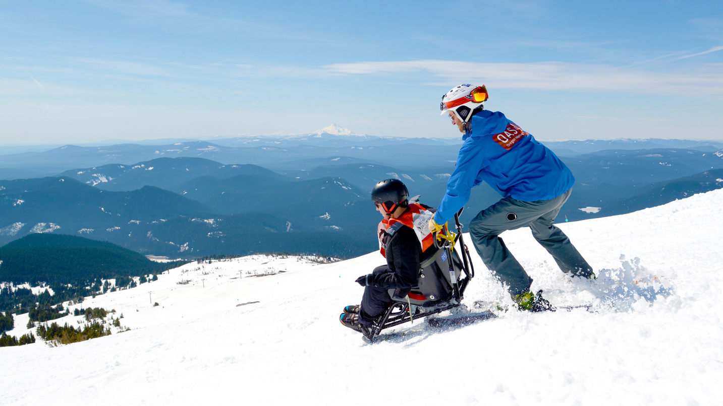 A guide skis downhill with a person in a mobility ski chair.