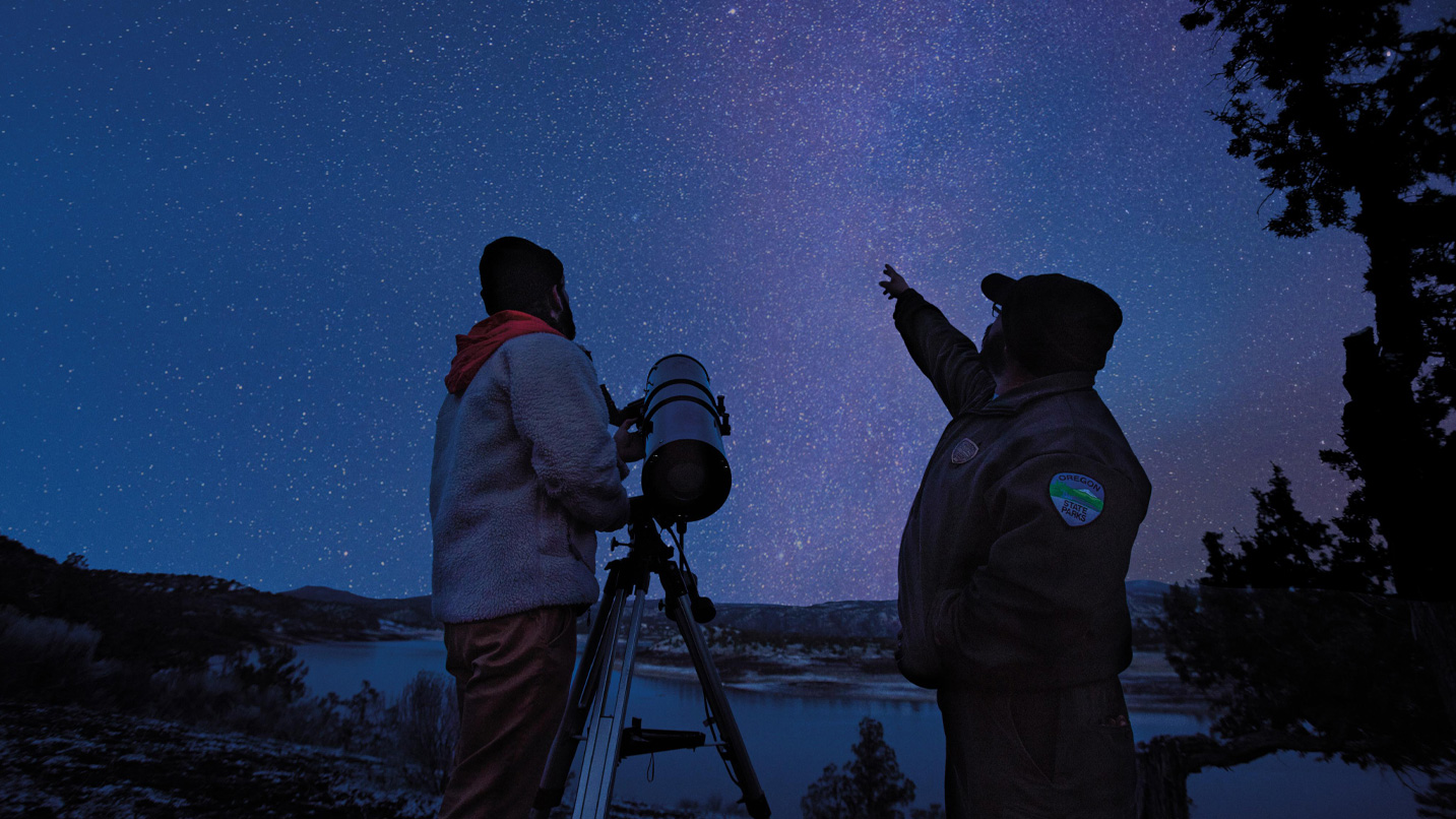 Park ranger and person with telescope looking up at a starry sky filled with stars.
