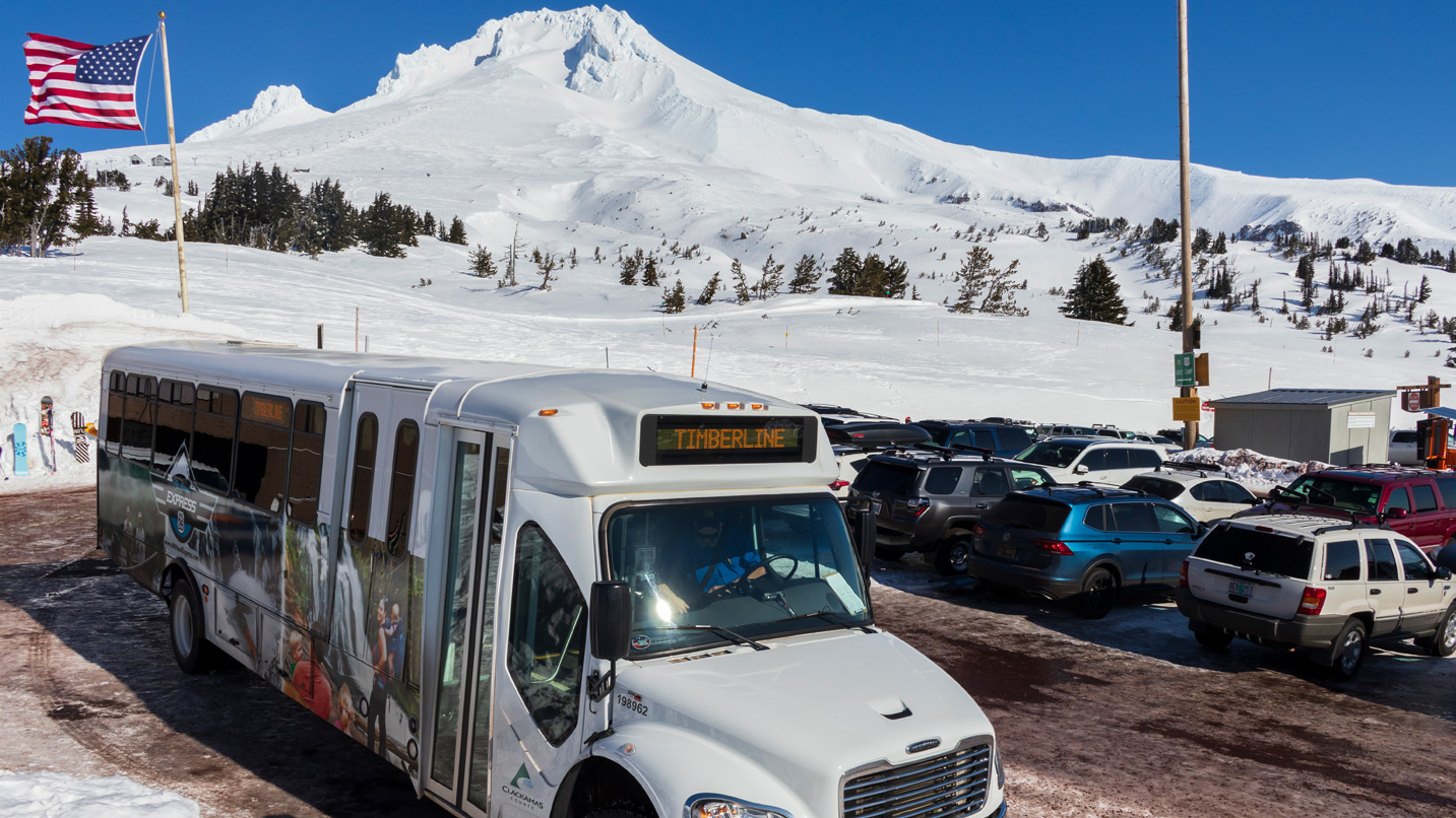 A shutttle buss parked in a parking lot with views of a snowy mountain at Timberline.