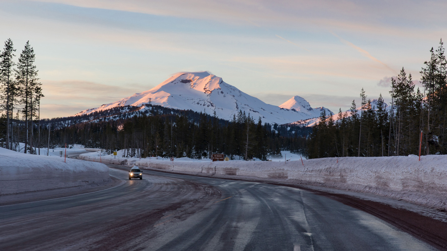 An icy highway road leading to a snowy mountain. A lone car drives on the highway.