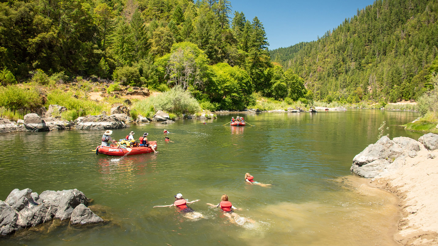 An idyllic scene of a calm river; groups of people are rafting and swimming in the river.