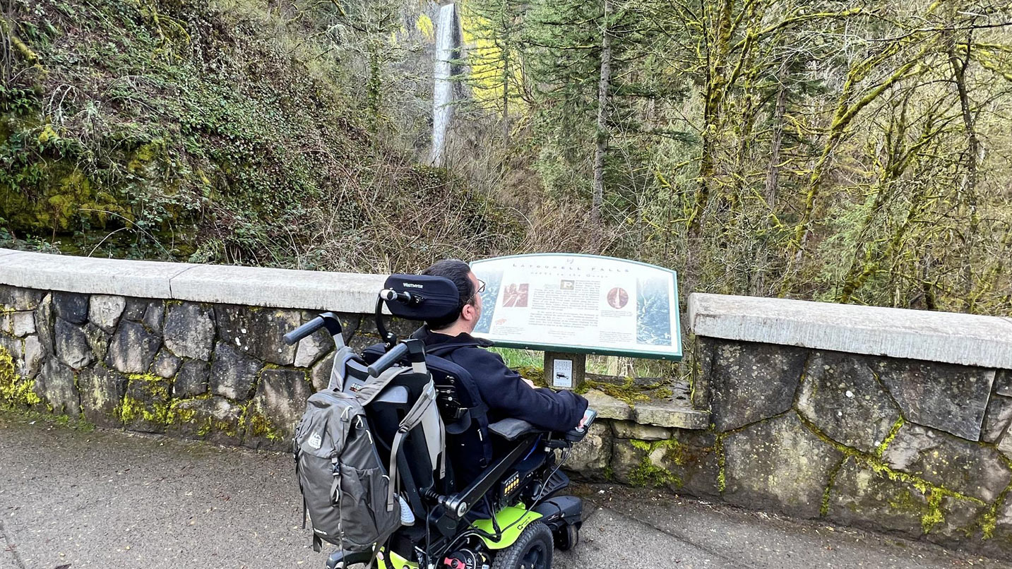 Man in a power wheelchair reads a sign on a trail.