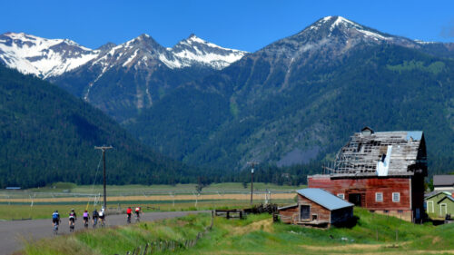 Cyclists on road next to old barn with snowcapped mountains ahead