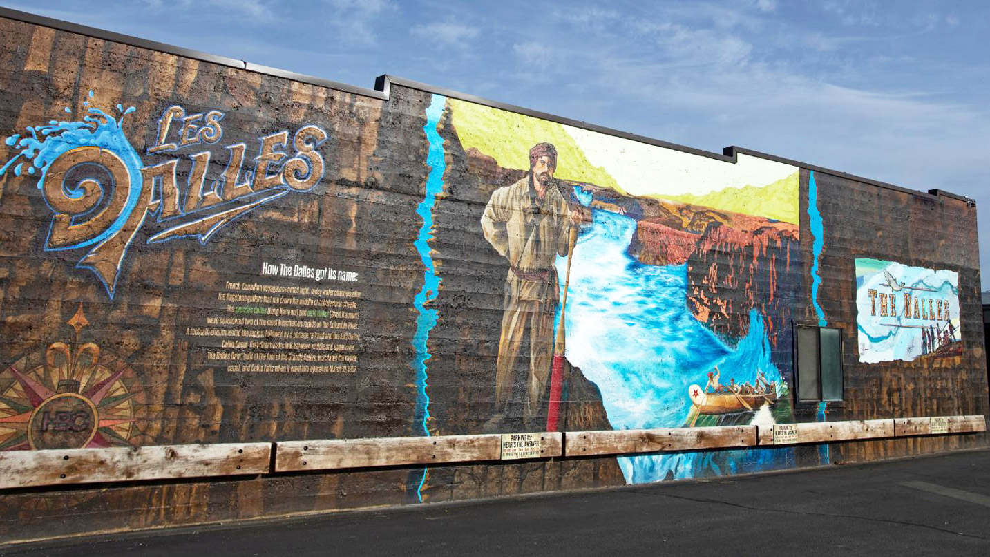 A mural depicting the Columbia River and a pioneer man with a paddle. Stylized font states "Les Dalles" and a small description of the The Dalles got its name, which is unreadable in the photo.