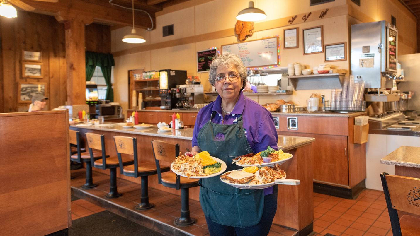 An employee holding 3 plates of food poses for a photo inside a diner.