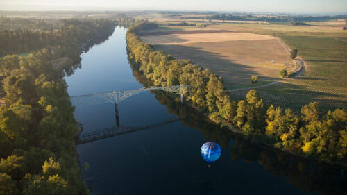 A hot air balloon floating above a river with a view of vineyards and a bridge