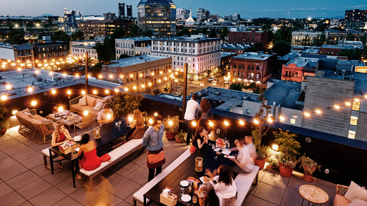 Rooftop bar space with people sitting at tables under lights with urban view