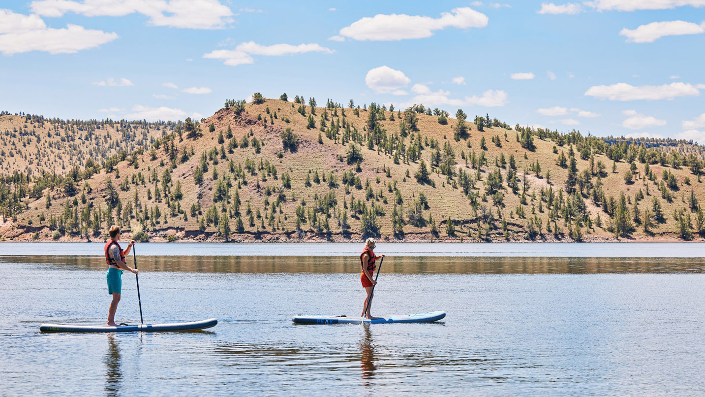 Two people stand paddleboarding on a calm lake, each wearing life jackets. A tree-covered hillside and a partly cloudy sky form the backdrop.