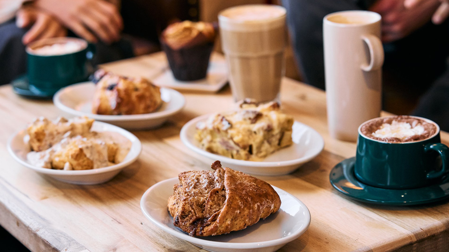 Pastries and coffee cups rest on a wooden table. Various baked goods sit on plates, accompanied by mugs and glasses of drinks, surrounded by people seated closely.
