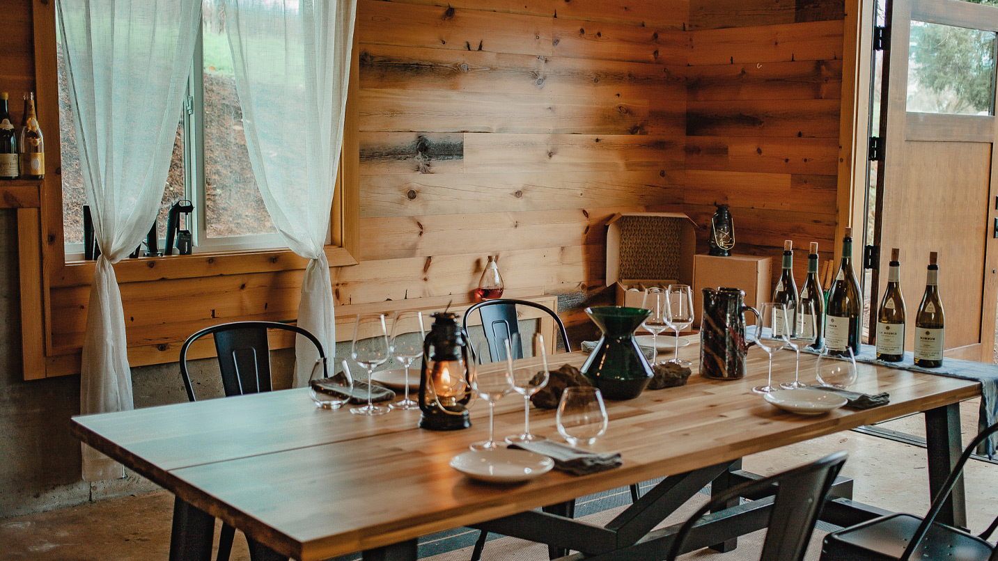A wooden dining table is set with wine glasses, plates, and lanterns inside a cozy room with wood-paneled walls, large window draped with curtains, and wine bottles displayed.