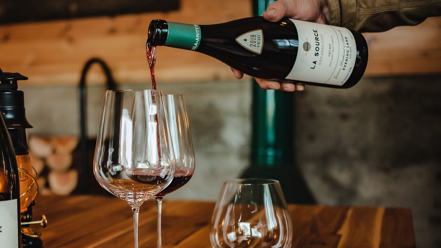 A hand pours red wine labeled "2019 Vintage La Source" into a glass on a wooden table in a cozy, rustic setting with blurred background elements.