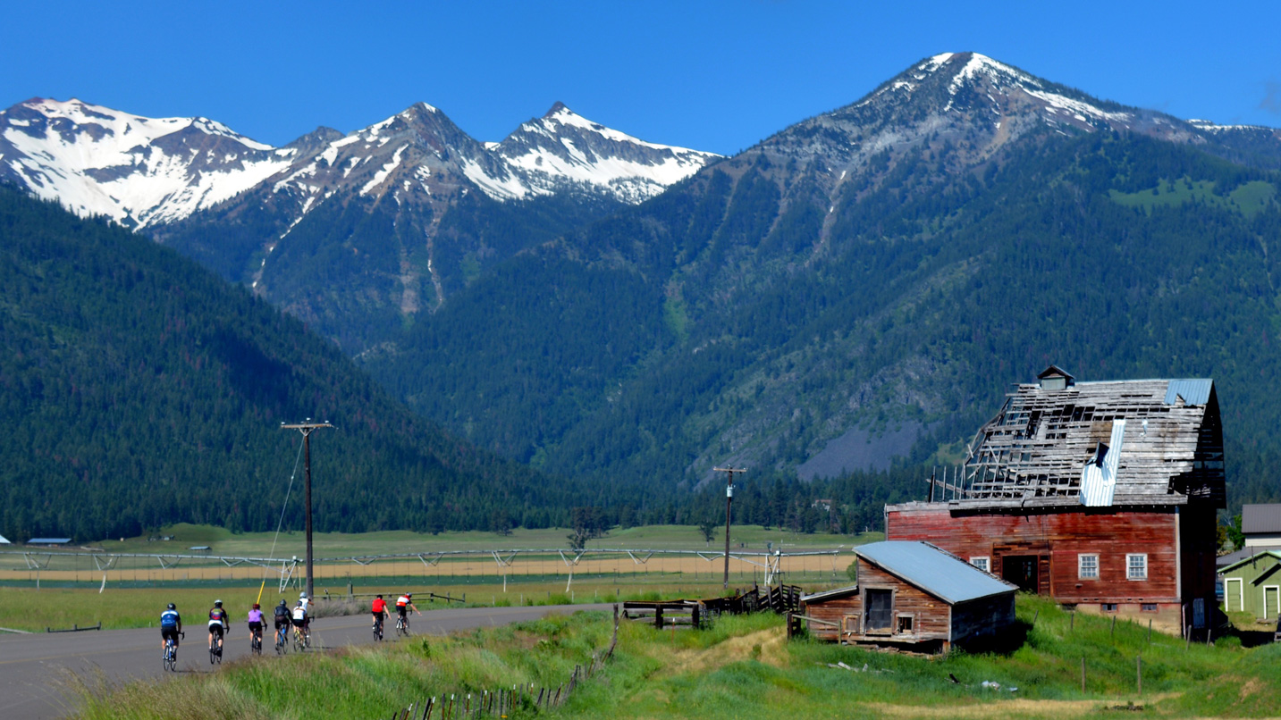 Cyclists in the foreground with red barns, grass and large snowcapped mountains in the background