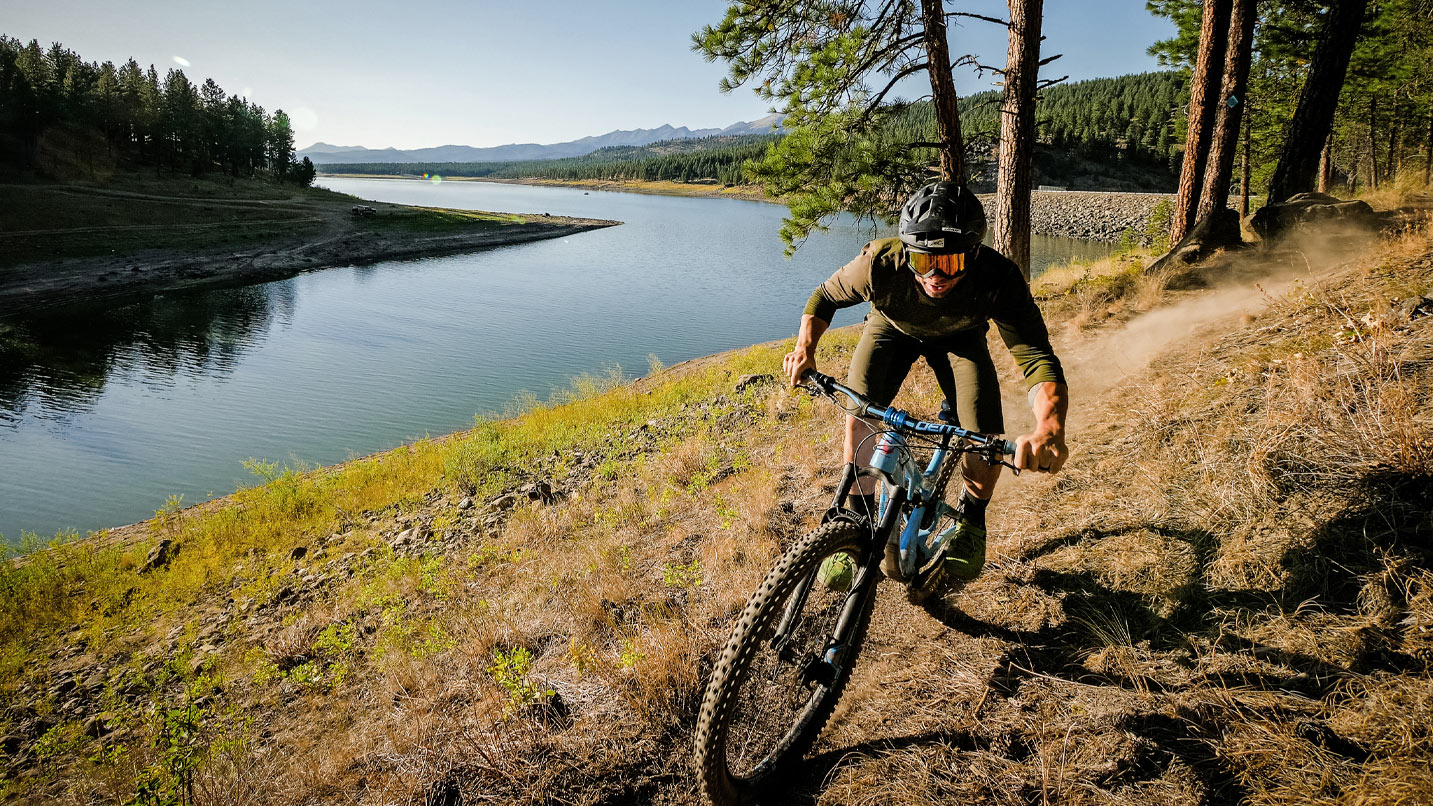 Man rides mountain bike on dirt trail next to lake and trees