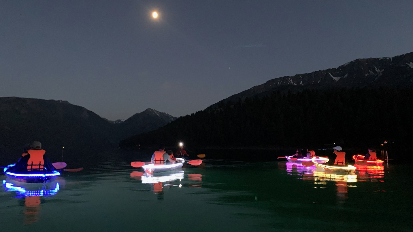 Several people on lit up kayaks paddle on a calm lake at night.