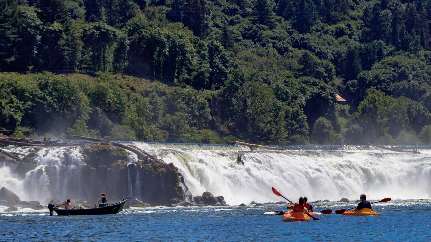 Several people kayaking on a river towards a view of rushing waterfalls.