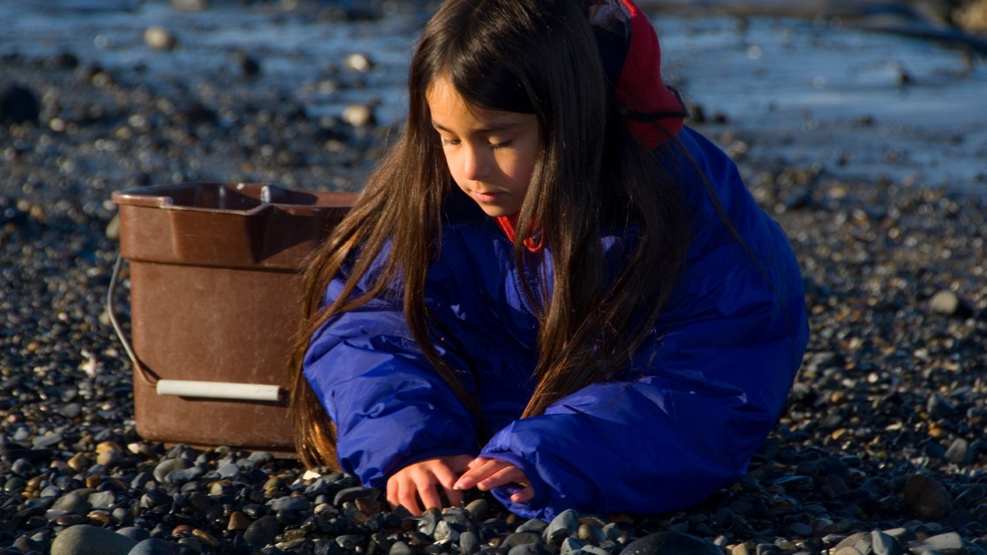 Girl plays with small rocks on beach with brown bucket beside her