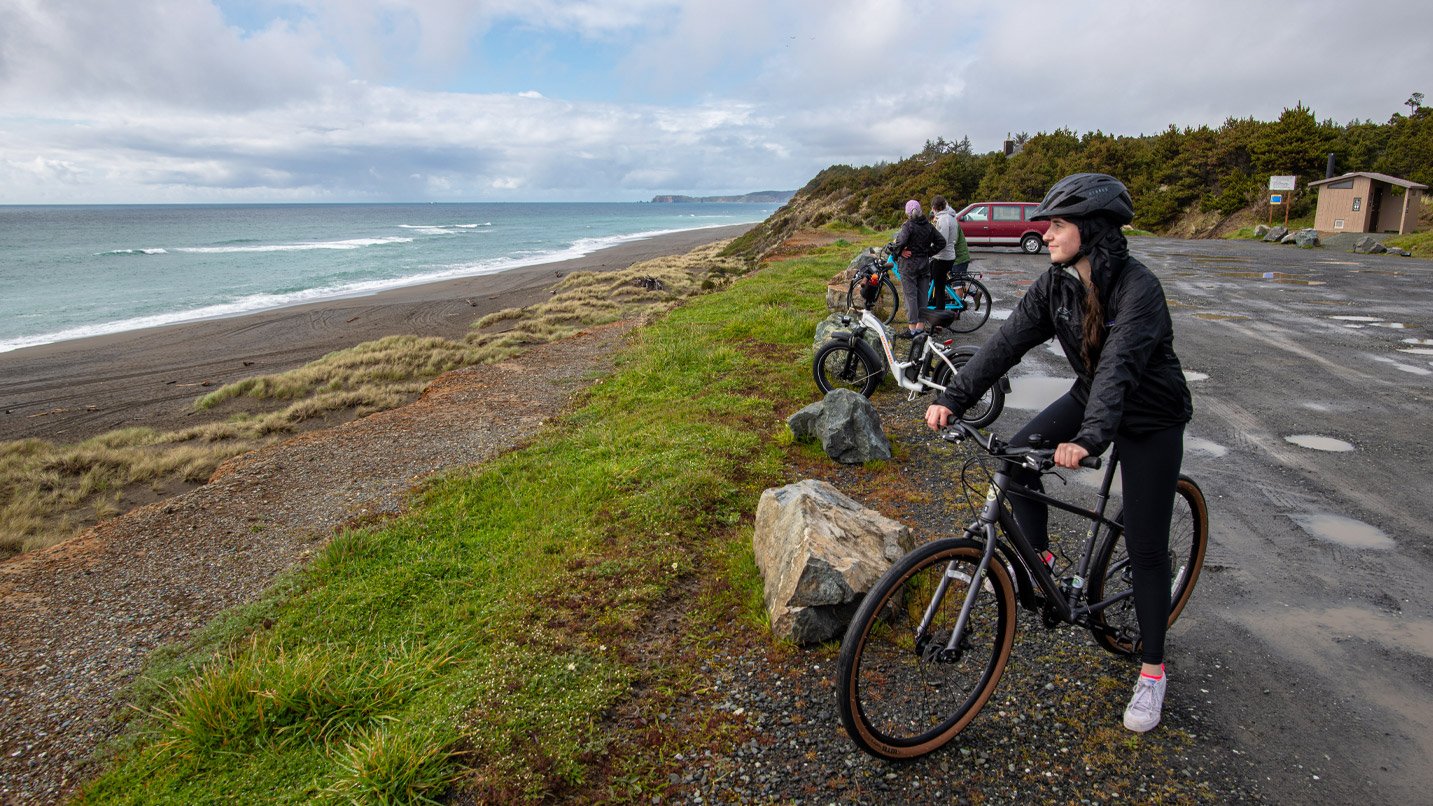 Woman in helmet stands with bike looking out at ocean