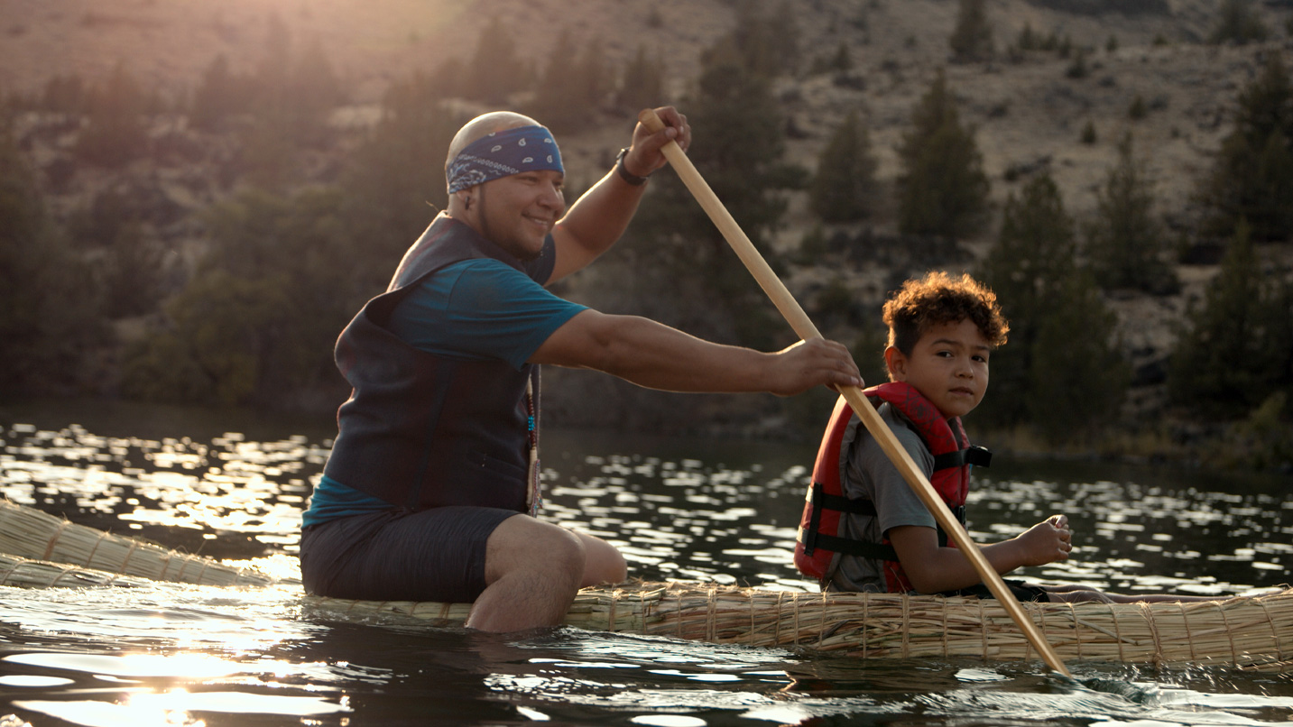 A man and child paddle a reed boat on a sunlit lake, surrounded by forested hills. The man is smiling, and the child is wearing a life vest.