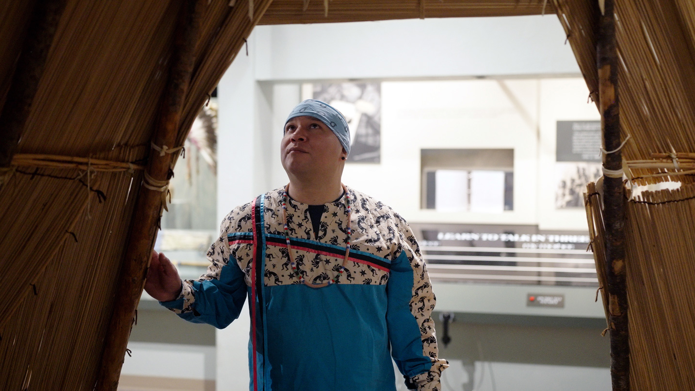 A person wearing a patterned outfit and headscarf stands inside a structure made of reeds and wood, looking upward, in a museum-like environment with informational displays in the background.