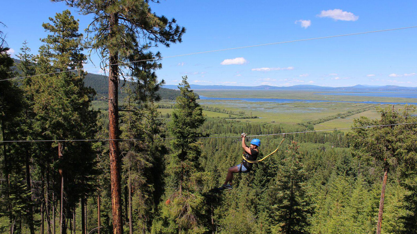 A person travels along a zipline with a lush green forest below them. The sky is clear with two small puffy clouds off into the distance.