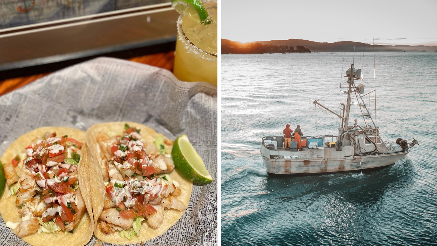 Tacos are filled with grilled fish, diced tomatoes, and drizzled with sauce, accompanied by a lime wedge on a napkin. A fishing boat sails on calm water at sunset.