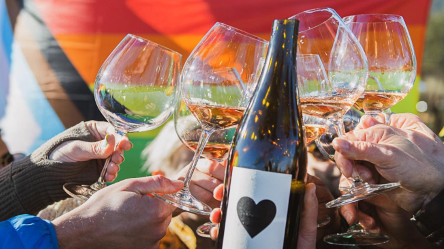 Close up of a wine bottle and several glasses of wine cheering with a pride flag in the background.