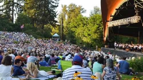 An orchestra performing on a stage in front of a large audience seated on festival grounds.